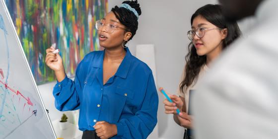 Two women standing at a whiteboard, one holding a marker and explaining a diagram while the other listens and takes notes during a collaborative discussion.
