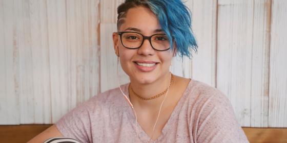 A person with short blue hair and glasses smiling at the camera while seated indoors, wearing earphones and a casual shirt.