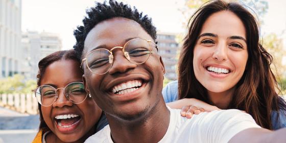 Three young adults smiling closely together while taking a selfie outdoors, with buildings visible in the background.