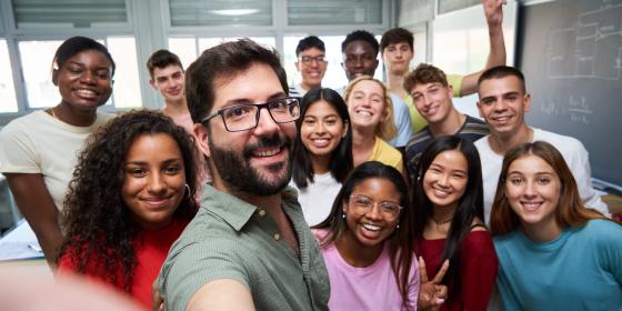 An adult taking a selfie with a large group of smiling students in a classroom, with desks and a chalkboard visible in the background.