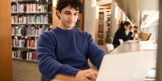 Student sitting at a long desk in a library, typing on a laptop, with bookshelves and other students studying in the background.