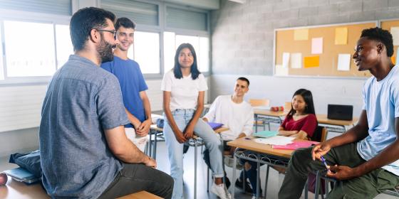 An adult facilitator sitting on a desk speaking with a small group of students seated around classroom desks during a group discussion.