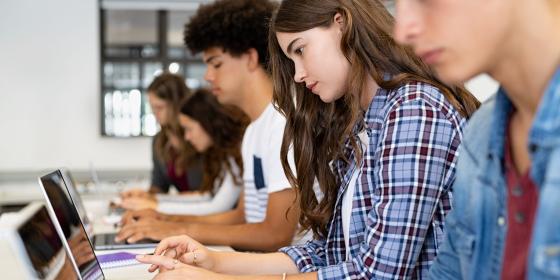 Students seated in a row at classroom desks, focused on typing on laptops during independent work time.