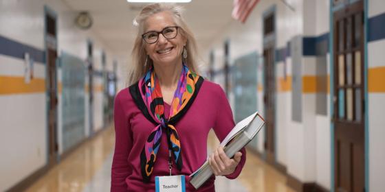 An adult educator standing in a school hallway, smiling and holding books, wearing a name badge that reads “Teacher.”
