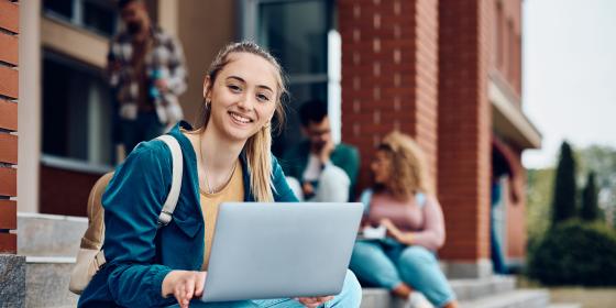A smiling student sitting on outdoor steps using a laptop, with other students gathered and talking in the background near a school building.