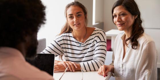 Two women seated at a table listening attentively to a person off camera during a meeting, with a laptop and documents on the table.